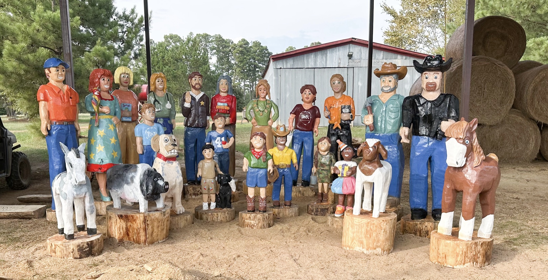 Hand carved and painted wooden characters from Teach BIG stories, including children, cowboys, a dog, a pig, a goat, and horses, standing on log stumps in front of a barn and hay bales.